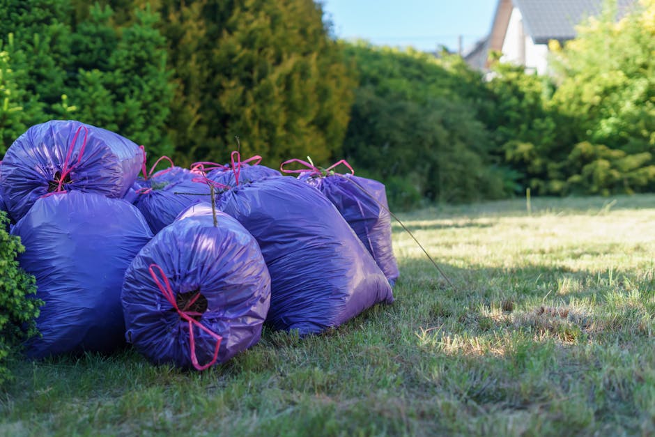 A group of several large, tightly packed purple plastic garbage bags filled with yard waste, resting on a grassy area next to a bush. The bags are secured with pink drawstrings and are positioned in a