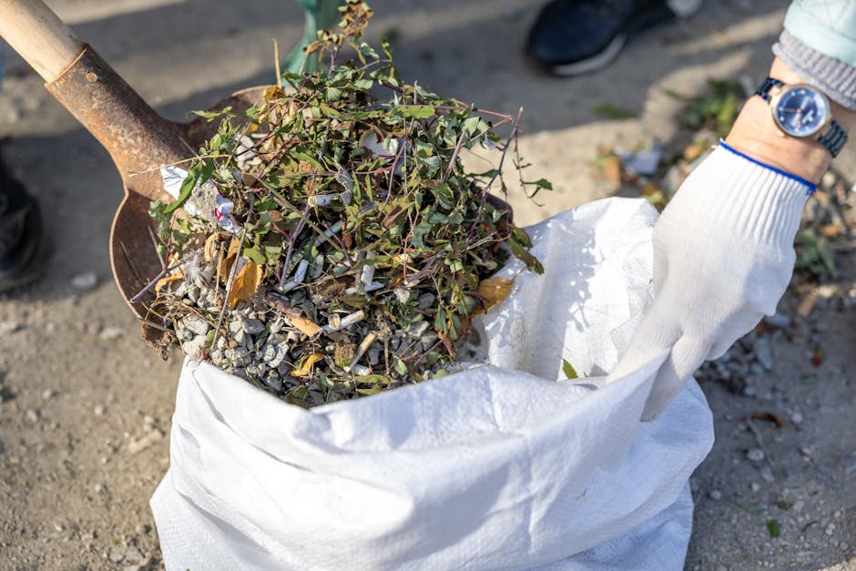 A group of several large, tightly packed purple plastic garbage bags filled with yard waste, resting on a grassy area next to a bush. The bags are secured with pink drawstrings and are positioned in a