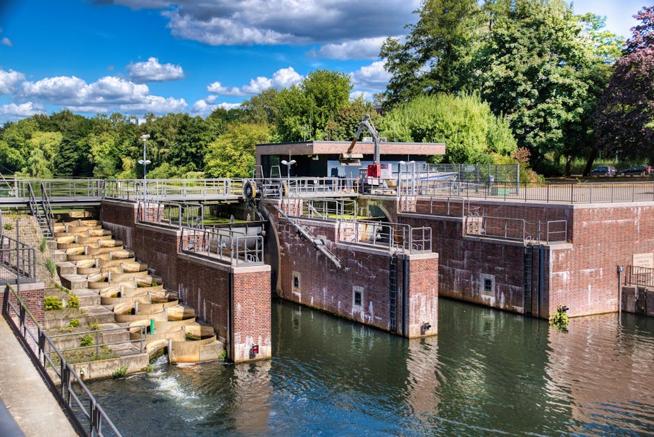 A brick wall and concrete embankment along a waterway, featuring metal railings on top and at the water level. The brick wall has areas of white staining and weathering, with some greenery and small p